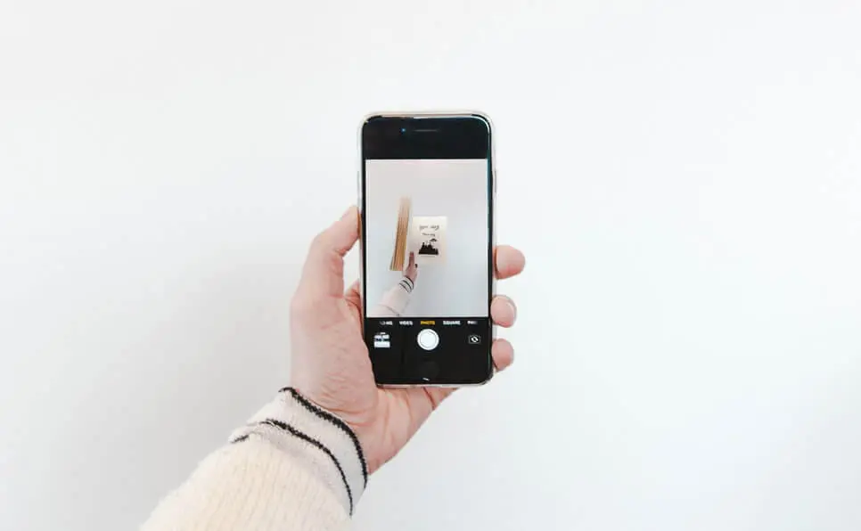 A person wearing a cream sweater holds a smartphone, taking a photo of a hand holding a small picture against a plain white wall. The phone screen displays the image being captured.