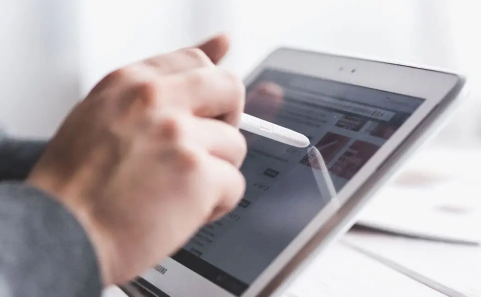 A person uses a stylus to interact with a white tablet, navigating a website on the device’s touchscreen. The background is blurred, drawing focus to the hand and tablet.