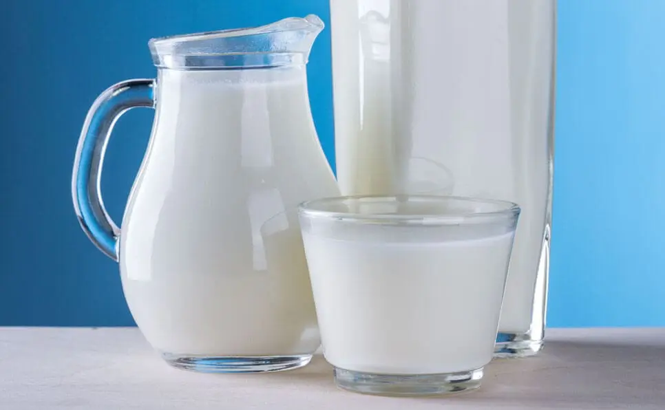 A glass pitcher, a glass, and a tall bottle, all filled with milk, are placed on a light-colored surface against a blue background.
