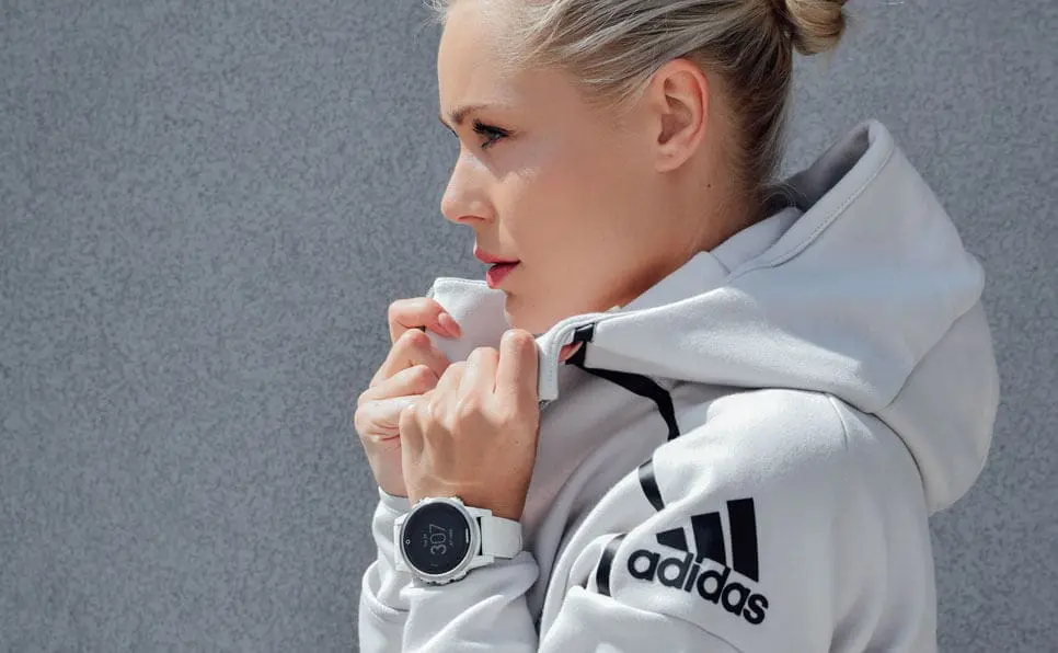 A woman in a light gray Adidas hoodie zips up her jacket while wearing a white smartwatch. She is standing in front of a textured gray wall, looking to the side with her hair pulled back.