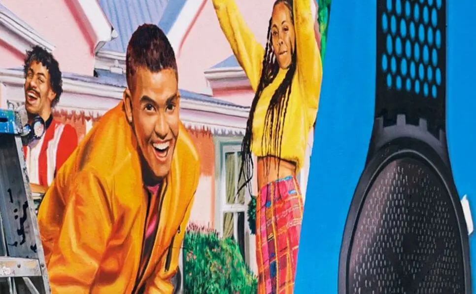 Three young people smile and dance outdoors, wearing colorful clothing. The background shows a pink house, greenery, and part of a large black and blue speaker on the right side of the image.