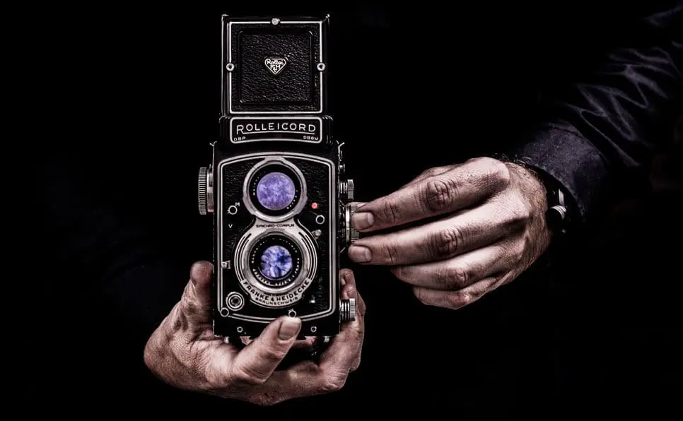 A person holds a vintage Rolleicord twin-lens reflex camera with both hands against a dark background, showcasing the cameras intricate details and lenses.