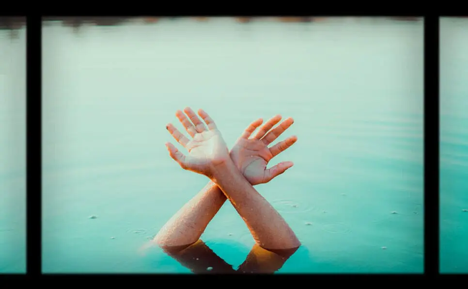 Two hands emerge from calm blue water, wrists crossed and palms open, creating an expressive gesture against a blurred background. The image is framed with black bars on the top, bottom, and sides.