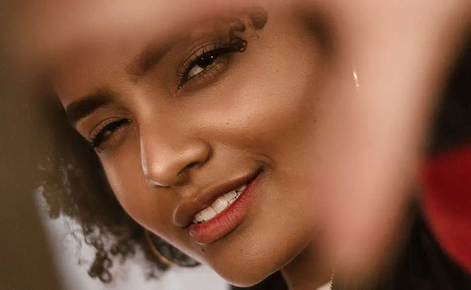 A person with curly hair and hoop earrings looks into the camera, partially framed by their hands. They have a slight smile and one eye squinting, creating a playful and confident expression.