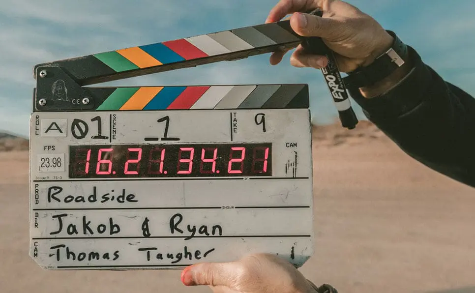 A person holds a film clapboard outdoors, ready to start a scene. The clapboard displays digital timecode and handwritten text, including Roadside, Jakob & Ryan, and Thomas Tangher. Desert landscape in background.